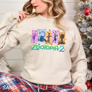 Puede incluir: Una sudadera de color arena con el texto "ZOOTOPIA 2" en turquesa e ilustraciones de personajes de dibujos animados. Las ilustraciones est&aacute;n enmarcadas en rect&aacute;ngulos de colores. La sudadera tiene mangas largas.