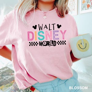 May include: A light pink short-sleeve t-shirt featuring the text WALT DISNEY WORLD in black, pink, blue, and purple lettering. The design includes two black Mickey Mouse ear silhouettes and a black and white checkered pattern border.