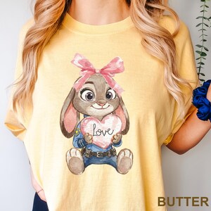 May include: Yellow t-shirt featuring a cartoon bunny holding a pink heart with the word "Love" on it. The bunny wears a pink bow and a police uniform. The word "BUTTER" is printed at the bottom right corner of the shirt.