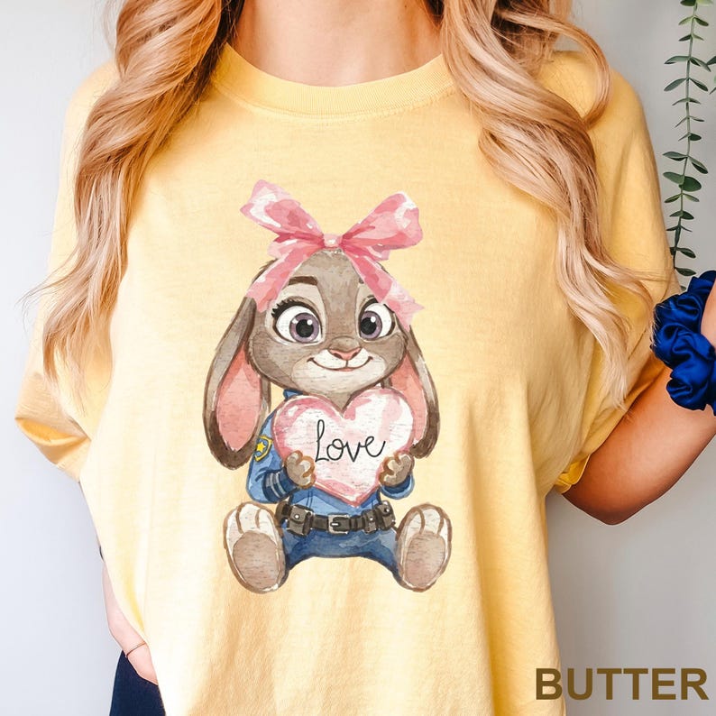 May include: Yellow t-shirt featuring a cartoon bunny holding a pink heart with the word "Love" on it. The bunny wears a pink bow and a police uniform. The word "BUTTER" is printed at the bottom right corner of the shirt.