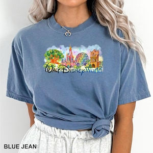 May include: A blue jean colored short-sleeve t-shirt featuring a colorful graphic print of the Walt Disney World theme park landmarks, including a castle, a geodesic sphere, and a tree, with the text Walt Disney World printed in blue.