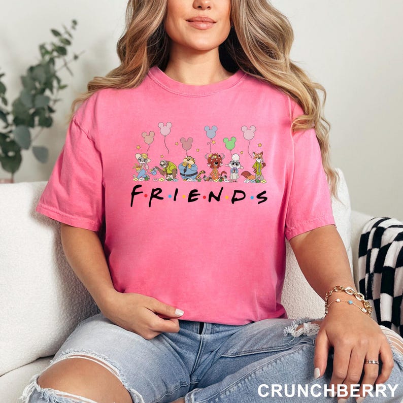 May include: A pink t-shirt with the word "FRIENDS" in black, featuring cartoon characters and Mickey Mouse-shaped balloons. The shirt is a solid color and has short sleeves.