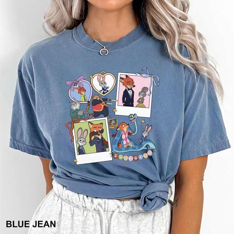 May include: Blue jean-coloured t-shirt with a vibrant graphic of Zootopia characters. The design incorporates polaroid-style frames and heart shapes, with various characters. The word "Zootopia" is also visible on the shirt.