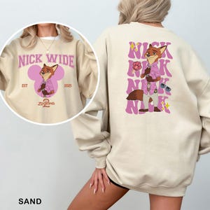 May include: Sand-colored sweatshirt featuring a cartoon fox character in a pink shirt and sunglasses. The front design includes the text "NICK WIDE" and "Zootopia 2". The back has a larger graphic of the fox with the name "NICK" repeated in a pink, wavy font.