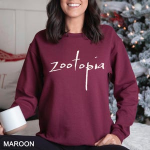 May include: A maroon-coloured sweatshirt featuring the word "Zootopia" in white script. The sweatshirt is a solid maroon colour. The word "MAROON" is printed in white at the bottom of the image.