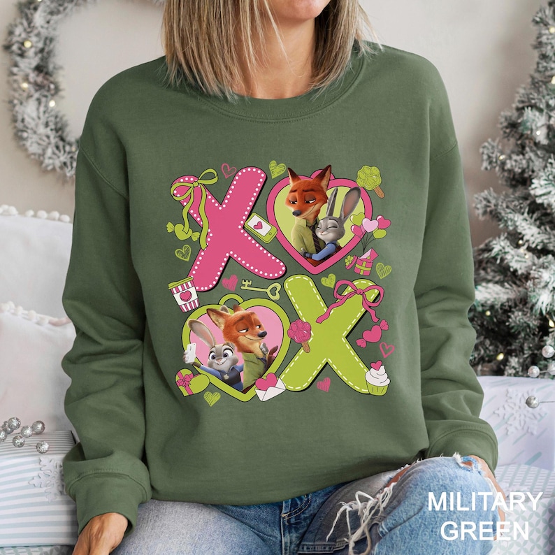 Puede incluir: Sudadera verde militar con un dise&ntilde;o gr&aacute;fico de los personajes Nick Wilde y Judy Hopps de Zootopia. El dise&ntilde;o incluye corazones rosas y verdes, las letras "X" y "O" y otros elementos tem&aacute;ticos de San Valent&iacute;n.