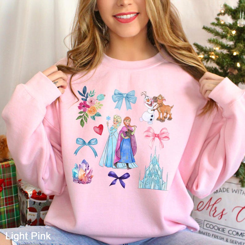 以下が含まれることがあります： 人気アニメのキャラクター、リボン、花、ハート、クリスタルを描いた水彩イラストが特徴のライトピンクのスウェットシャツ。デザインにはエルサ、アナ、オラフ、お城が含まれています。「Light Pink」の文字が見えます。