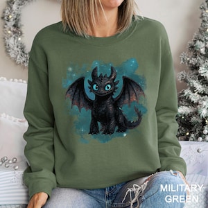 May include: Military green sweatshirt featuring a black dragon with blue eyes and wings. The dragon is set against a teal and blue background with stars. The text "MILITARY GREEN" is at the bottom right.