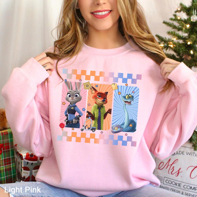 Puede incluir: Sudadera rosa claro con un dise&ntilde;o de tres personajes de dibujos animados: un conejo con uniforme de polic&iacute;a, un zorro con chaqueta verde y una serpiente. El dise&ntilde;o incluye un patr&oacute;n de cuadros y estrellas. El texto "Light Pink" es visible.