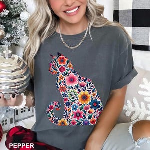 May include: A gray t-shirt featuring a floral cat design. The cat silhouette is filled with colorful flowers in shades of pink, orange, yellow, and blue. The word "PEPPER" is printed at the bottom.