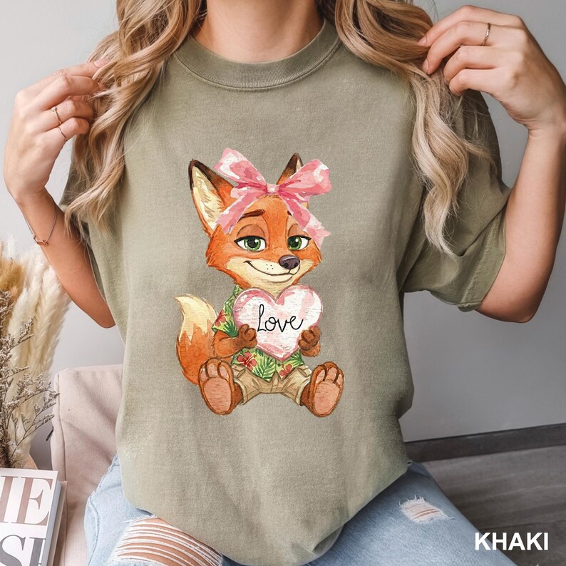 May include: Khaki-colored t-shirt featuring a cartoon fox holding a heart-shaped sign that says "Love." The fox has orange fur, a pink bow, and is wearing a Hawaiian shirt. The word "KHAKI" is printed in white at the bottom.