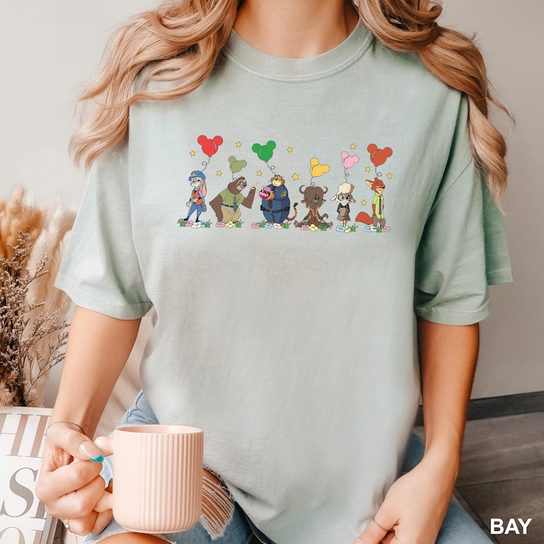 May include: A light green t-shirt featuring a whimsical cartoon design of various animal characters holding heart-shaped balloons. The characters are in a row, each with a unique outfit and expression. The balloons are in red, green, yellow, and pink.