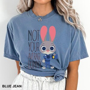 May include: Blue jean coloured t-shirt with a cartoon bunny design. The bunny has pink ears and the text reads "NOT YOUR AVERAGE BUNNY". The bunny is wearing a blue uniform and holding a bag and a lantern. The shirt is a casual style.