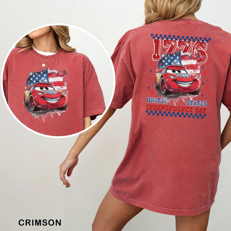 May include: A crimson red t-shirt featuring a graphic of a red race car in front of an American flag. The back design includes the text 1776 and United States Independence Day in blue and red lettering with a checkered border pattern.