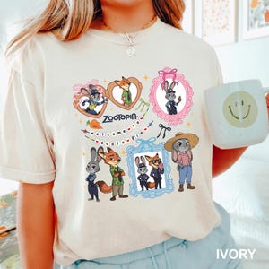 Op de afbeelding: Ivoor T-shirt met cartoonfiguren uit Zootopia. Het ontwerp bevat ingelijste afbeeldingen van een konijn en een vos, met de tekst "Zootopia" en "Welcome to Zootopia". Een witte mok met een smiley is ook zichtbaar.