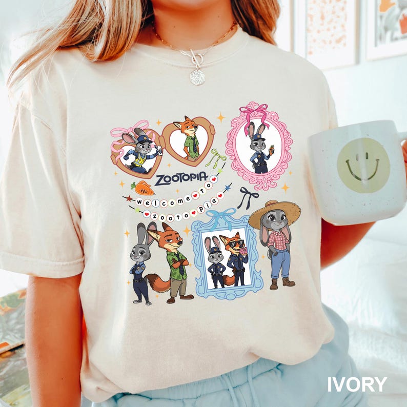 Op de afbeelding: Ivoor T-shirt met cartoonfiguren uit Zootopia. Het ontwerp bevat ingelijste afbeeldingen van een konijn en een vos, met de tekst "Zootopia" en "Welcome to Zootopia". Een witte mok met een smiley is ook zichtbaar.