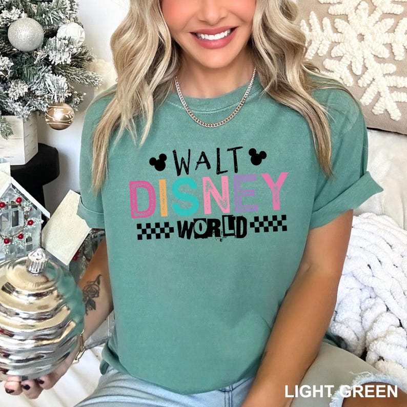 May include: A sage green short-sleeve t-shirt featuring the text WALT DISNEY WORLD in a mix of black, pink, and teal block lettering. The design includes two small black Mickey Mouse ear silhouettes and black checkered patterns on the sides.