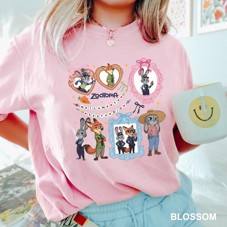 Op de afbeelding: Roze T-shirt met cartoonfiguren uit Zootopia. Het ontwerp bevat ingelijste afbeeldingen van een vos en een konijn, met de tekst "Zootopia" en "welcome to Zootopia". Het shirt heeft een ronde hals en korte mouwen.