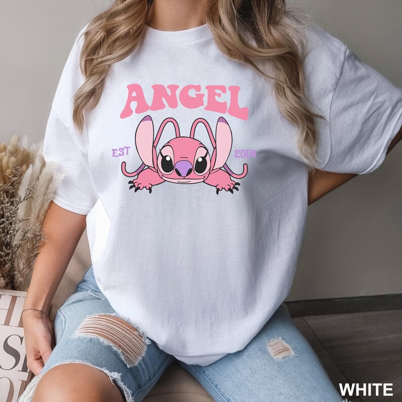 K&ouml;nnte beinhalten: Wei&szlig;es T-Shirt mit einer rosa Cartoonfigur und dem Wort "ANGEL" dar&uuml;ber. Die Figur hat gro&szlig;e Ohren und liegt. Der Text "EST 2000" befindet sich ebenfalls auf dem T-Shirt.
