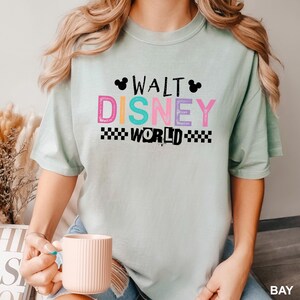 May include: A light green short-sleeve t-shirt featuring the text WALT DISNEY WORLD in colorful block letters. The design includes two small black Mickey Mouse head silhouettes and a black and white checkered pattern below the word WORLD.