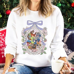 May include: White sweatshirt featuring a colorful cartoon illustration of various animal characters within an oval frame, surrounded by flowers and a light purple bow. The design includes characters from a popular animated movie.