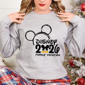 以下が含まれることがあります： 黒いミッキーマウスの頭のデザインと「DISNEY 2026 FAMILY VACATION」の文字が入った、ライトグレーのスウェットシャツ。スウェットシャツはスポーツグレーの色です。