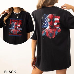 Może przedstawiać: Czarny t-shirt z grafiką czerwono-niebieskiej maski superbohatera na tle postarzałej amerykańskiej flagi. Z przodu znajduje się napis 1776, United States oraz Independence Day, a z tyłu umieszczono większą wersję tej samej grafiki.