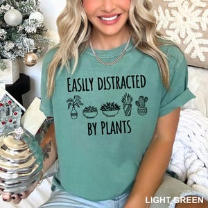 May include: A light green short-sleeve t-shirt featuring black text that reads EASILY DISTRACTED BY PLANTS above and below a line of five black line-art illustrations of various potted plants, including succulents and a cactus.