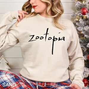 May include: A sand-coloured sweatshirt featuring the word "Zootopia" in black script. The long-sleeved crew neck top is worn by a person. The person is wearing plaid pyjama bottoms. A Christmas tree is in the background.