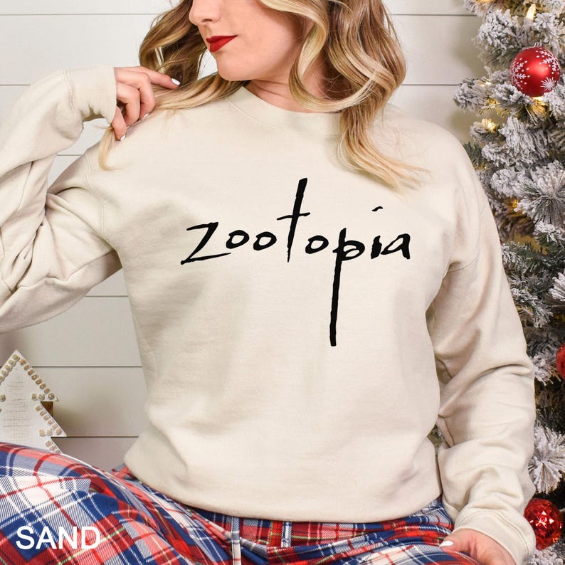May include: A sand-coloured sweatshirt featuring the word "Zootopia" in black script. The long-sleeved crew neck top is worn by a person. The person is wearing plaid pyjama bottoms. A Christmas tree is in the background.