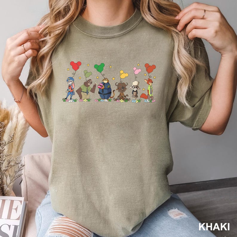 May include: Khaki-colored t-shirt featuring a whimsical illustration of cartoon characters holding heart-shaped balloons. The design includes characters in various colors, with the word "KHAKI" printed at the bottom right.