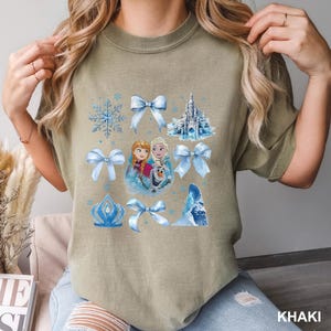 May include: Khaki-colored t-shirt featuring a collage of blue Frozen-themed graphics. The design includes snowflakes, bows, a castle, and images of Anna, Elsa, and Olaf. The word "KHAKI" is printed in the bottom right corner.