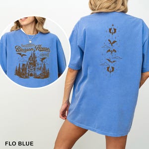May include: Flo blue t-shirt with a brown graphic of a castle, dragons, and text that reads "Riorrson House" on the front. The back of the shirt features a vertical design of dragons and clouds.