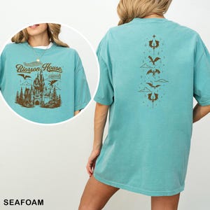 May include: Seafoam green t-shirt with a brown graphic on the front featuring a castle, dragons, and the text "Riorrson House". The back of the shirt has a vertical design of dragons and clouds. The shirt is made of a soft material.
