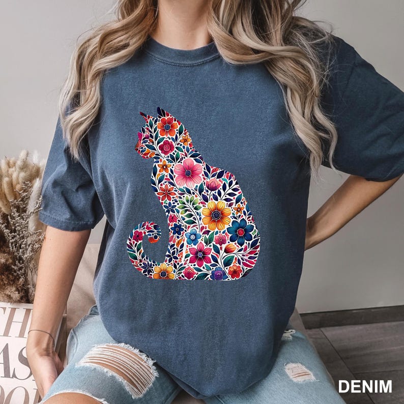 May include: A denim blue t-shirt featuring a floral cat design. The cat silhouette is filled with colorful flowers in shades of pink, yellow, blue, and orange. The word "DENIM" is printed in white at the bottom right.