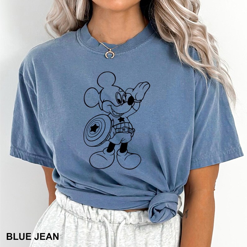 May include: Blue jean colored t-shirt featuring a black graphic of Mickey Mouse as Captain America. Mickey is holding a shield with a star and wearing a Captain America belt. The shirt has a crew neck and short sleeves.