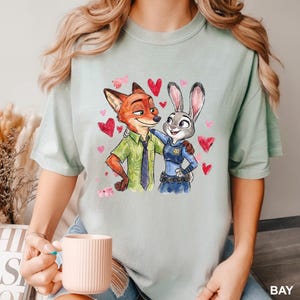 Może przedstawiać: Jasnozielona koszulka z akwarelową ilustracją Nicka Wilde'a i Judy Hopps z Zootopii, otoczona czerwonymi sercami. Nick ma na sobie zieloną koszulę i krawat, a Judy mundur policjantki.