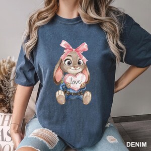 May include: Denim blue t-shirt featuring a cartoon bunny holding a heart with the word "Love". The bunny has a pink bow and is wearing a blue uniform. The shirt is a comfortable, casual style.