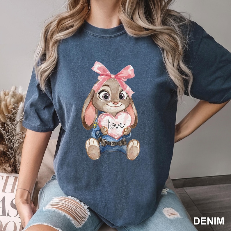 May include: Denim blue t-shirt featuring a cartoon bunny holding a heart with the word "Love". The bunny has a pink bow and is wearing a blue uniform. The shirt is a comfortable, casual style.