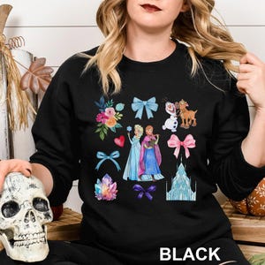 以下が含まれることがあります： 人気アニメ映画のキャラクター、リボン、花、クリスタルをあしらった、カラフルな水彩イラストが特徴の黒いクルーネックのスウェットシャツ。「BLACK」の文字が白でプリントされています。