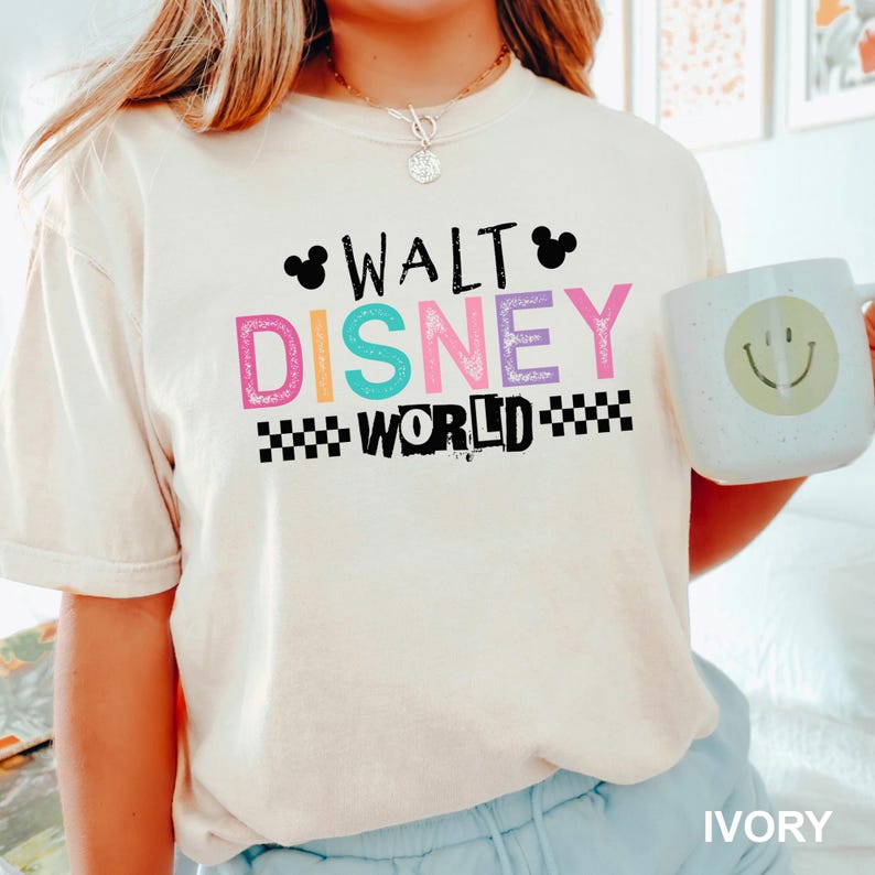 May include: An ivory short-sleeve t-shirt featuring the text WALT DISNEY WORLD in black, pink, teal, and purple. The design includes two black Mickey Mouse ear silhouettes and a black and white checkered pattern. A light-colored mug with a smiley face.