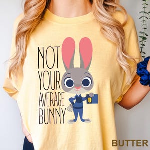 May include: Yellow t-shirt featuring the text "NOT YOUR AVERAGE BUNNY" and a cartoon bunny character. The bunny is grey with pink ears, wearing a blue uniform, and holding a blue briefcase. The word "BUTTER" is at the bottom right.