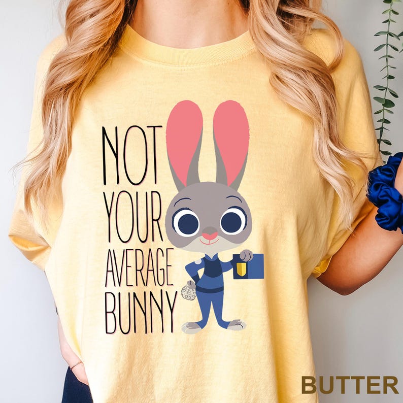 May include: Yellow t-shirt featuring the text "NOT YOUR AVERAGE BUNNY" and a cartoon bunny character. The bunny is grey with pink ears, wearing a blue uniform, and holding a blue briefcase. The word "BUTTER" is at the bottom right.