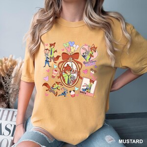 Op de afbeelding: Mosterdkleurig T-shirt met cartoonfiguren uit Zootopia. Het ontwerp bevat Nick Wilde en Judy Hopps, samen met de tekst "Best Friends Forever" en "Bestie". Het shirt heeft een ronde hals en korte mouwen.