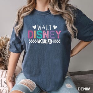May include: A dark blue short-sleeve t-shirt featuring the text WALT DISNEY WORLD in a multi-colored, distressed font. The design includes small white Mickey Mouse head icons above the text and white checkered patterns on the sides of the word WORLD.