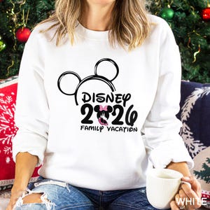 以下が含まれることがあります： ミッキーマウスの黒い頭の輪郭と「DISNEY 2026 FAMILY VACATION」の文字が入った白いスウェットシャツ。デザインにはピンクのリボンが含まれています。スウェットシャツはクルーネックで長袖です。