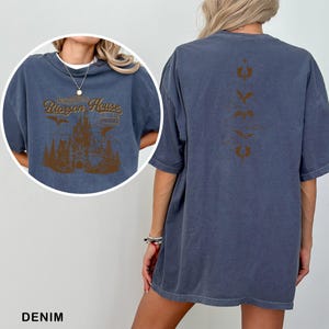 May include: Denim blue t-shirt with a brown graphic on the front featuring the text "Rioron House" above a castle illustration. The back of the shirt has a vertical brown design. The shirt is a comfortable, oversized fit.