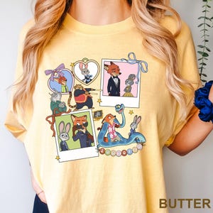 May include: Butter-yellow t-shirt with cartoon characters from Zootopia. The design includes character portraits in polaroid-style frames, with the word "Zootopia" in a circular pattern. The shirt has a crew neck and short sleeves.