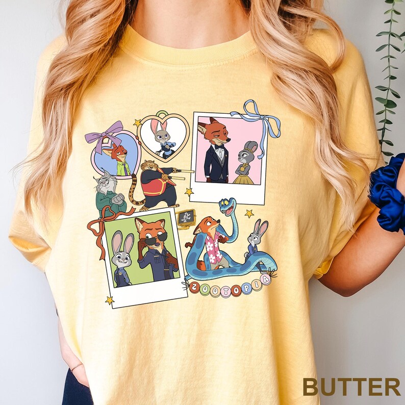 May include: Butter-yellow t-shirt with cartoon characters from Zootopia. The design includes character portraits in polaroid-style frames, with the word "Zootopia" in a circular pattern. The shirt has a crew neck and short sleeves.