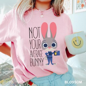 May include: A pale pink t-shirt featuring the phrase "NOT YOUR AVERAGE BUNNY" and a cartoon depiction of a bunny in a police uniform. The bunny has large ears and is holding a badge and a flashlight. A white mug with a yellow smiley face is also visible.